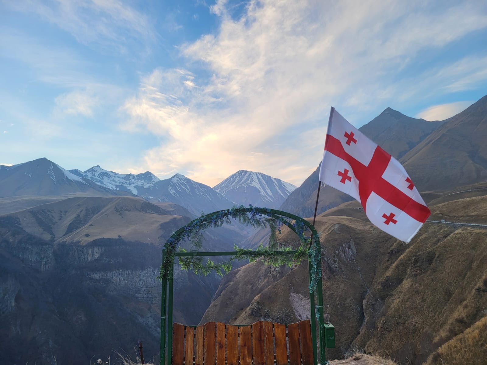 A flower-wound arch and the Georgian flag on a high ridge above a valley, with snow-capped peaks beyond